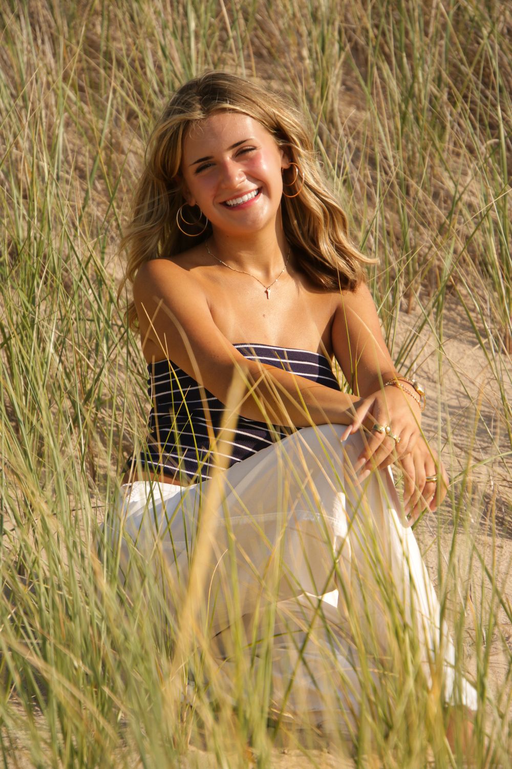 Woman in striped dress poses joyfully amid tall beach grass in summer sunlight.