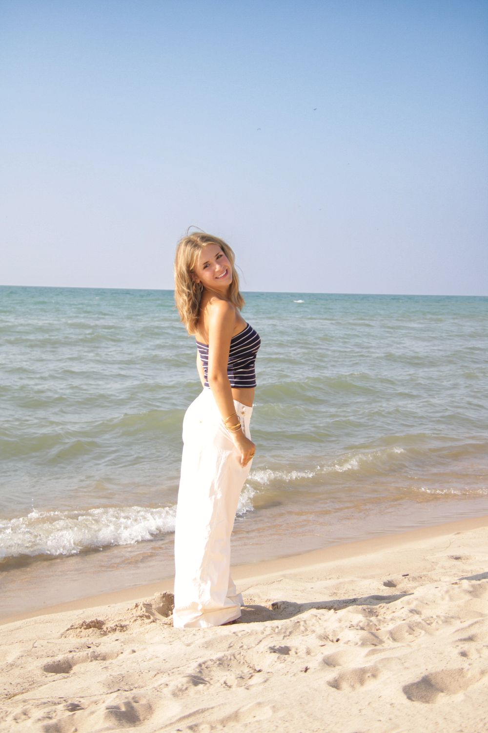 Person in striped top and white pants posing on sandy beach with ocean waves in background.