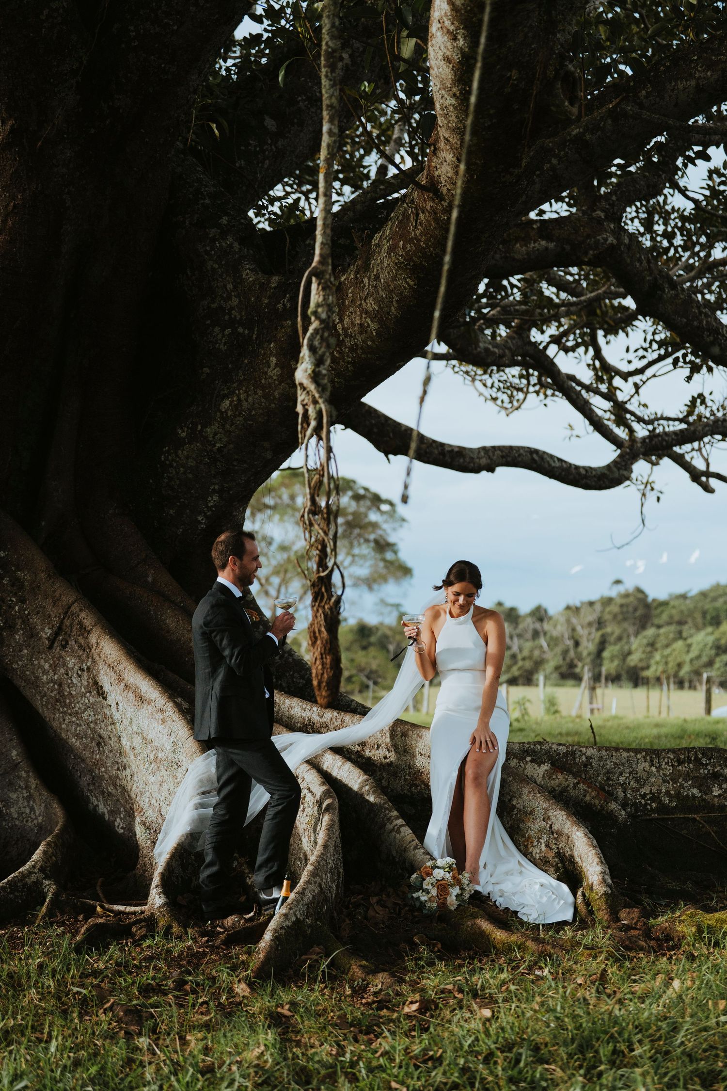 A romantic couple poses together under a majestic old tree in a scenic outdoor setting wearing wedding attire.