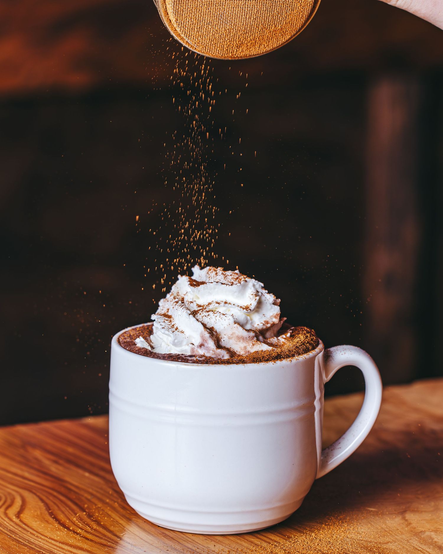 A cup of hot cocoa topped with whipped cream and chocolate powder being sprinkled on wooden table.