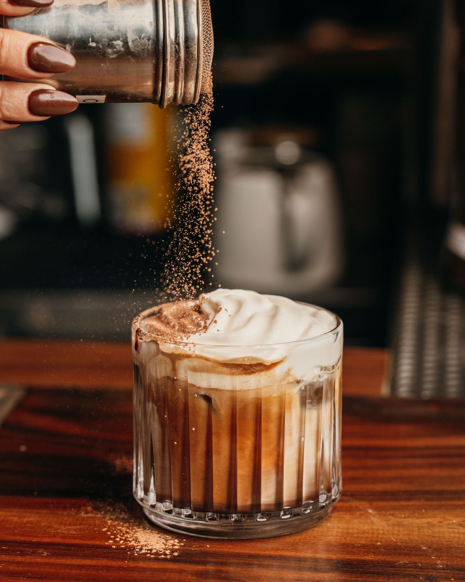 Cocoa powder being sprinkled onto a creamy drink with layered colors in a clear glass on a wooden surface.