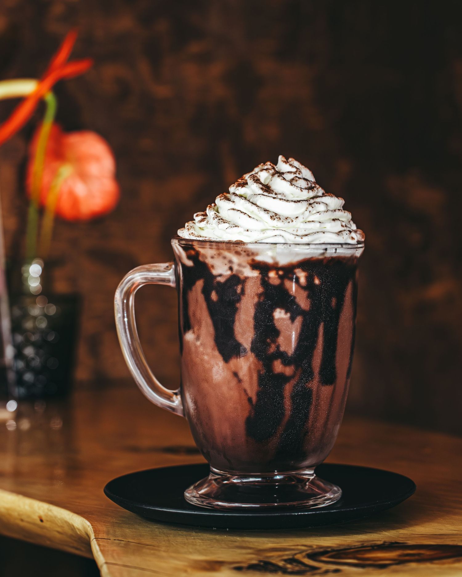 Hot chocolate in clear glass mug topped with whipped cream and chocolate drizzle on dark wooden surface.