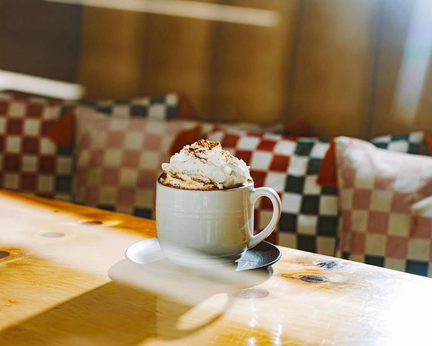 A cup of cappuccino with delicate latte art featuring a heart design sits on a white saucer with a silver spoon.