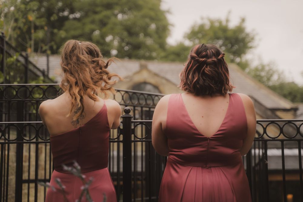 Bridesmaids in burgundy dresses line up against iron railing at outdoor venue showing matching backs of gowns.