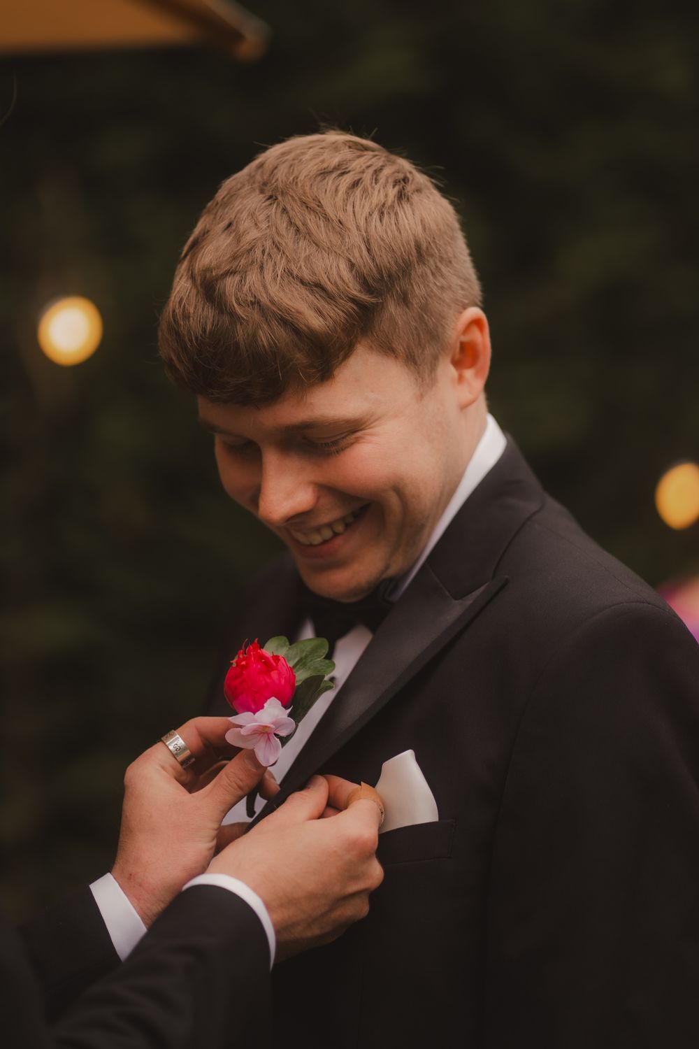 Groomsman in black tuxedo adjusts a pink rose boutonniere at an evening wedding ceremony.