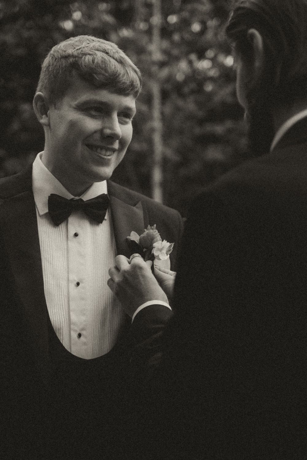 A young person in a black tuxedo and bow tie smiles while wearing a boutonniere at a formal event.
