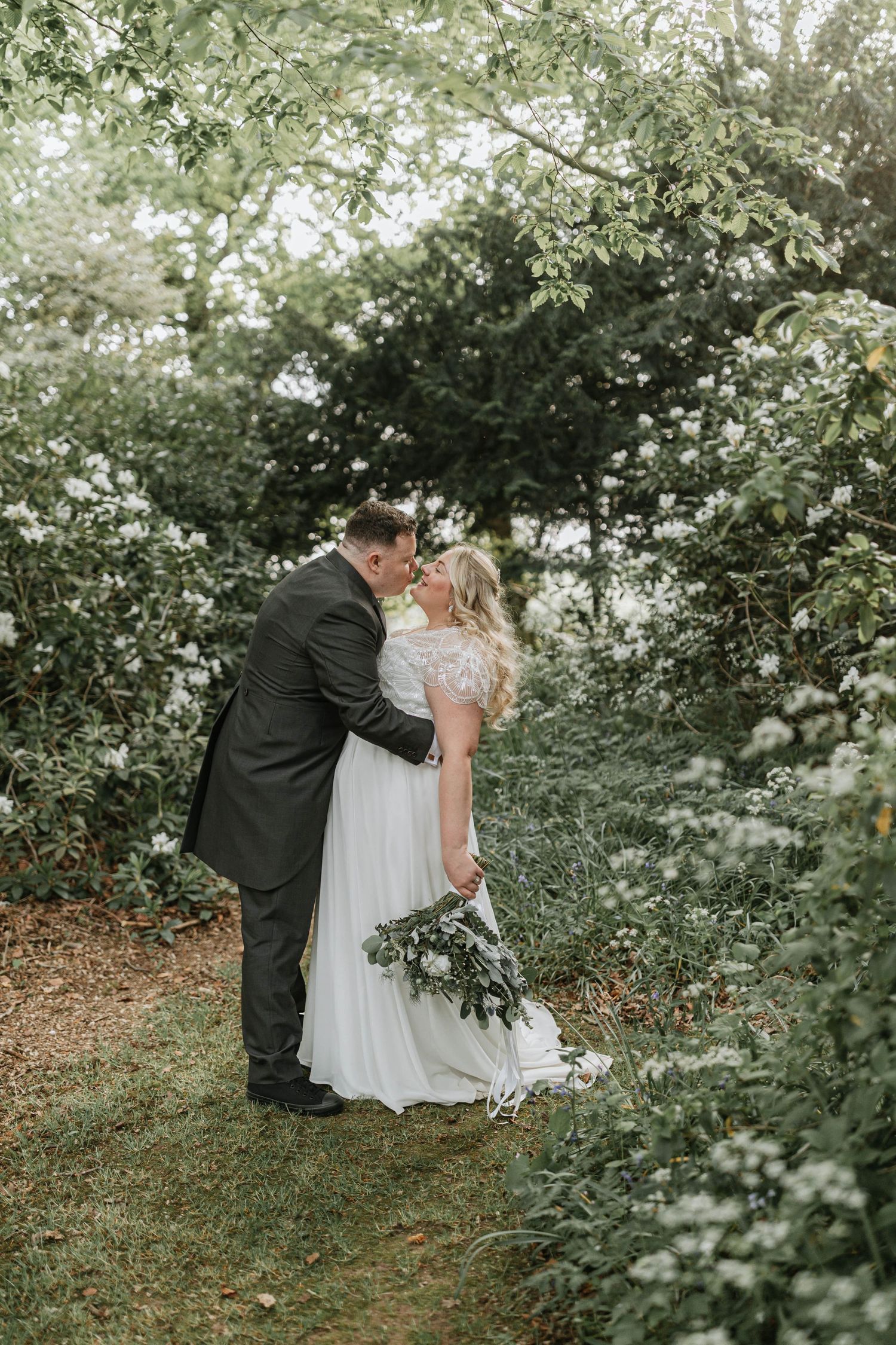 A bride in white and groom in black embrace romantically in a lush garden setting with flowering trees.