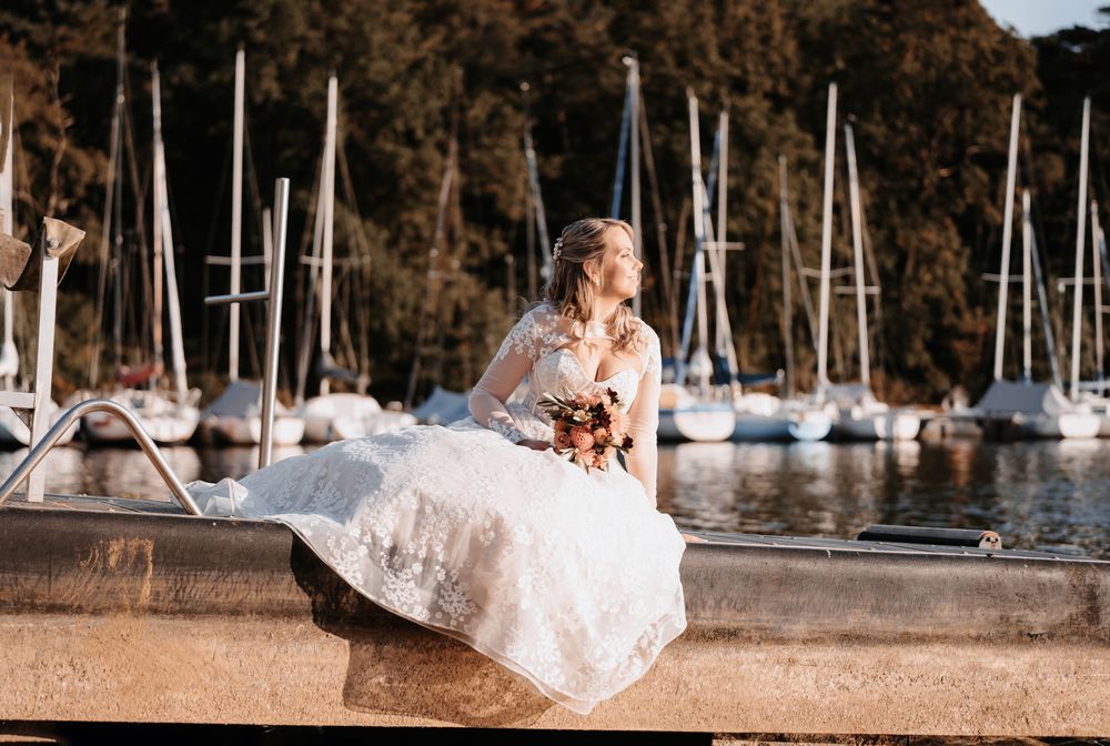 Einzelaufnahme der Braut am Steg, Kleid elegant drapiert, Beine hochgelegt, sie blickt in die Sonne, Segelschiffe unscharf im Hintergrund