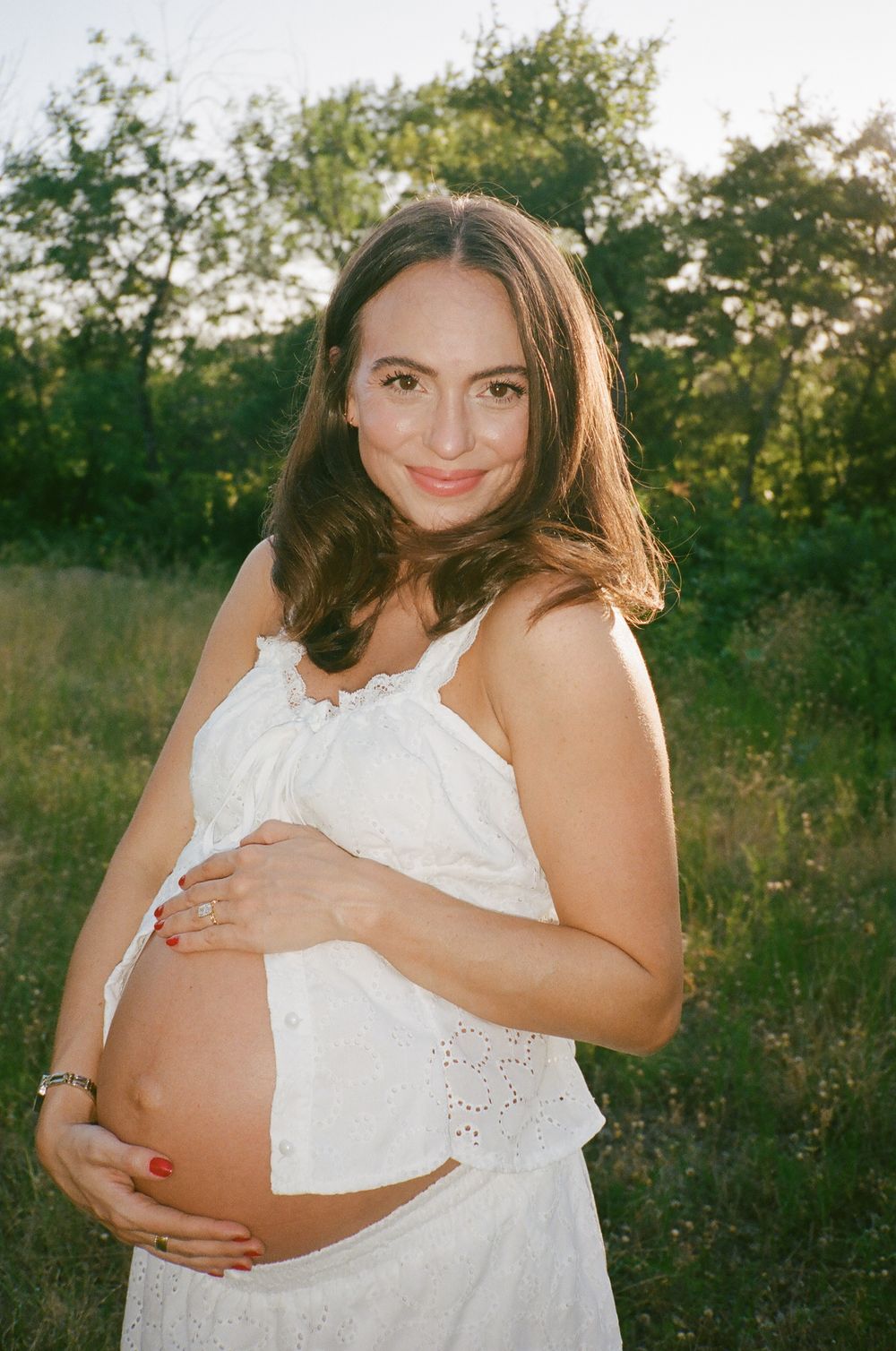 Pregnant woman in white dress cradling baby bump outdoors in sunlit green field, smiling warmly.