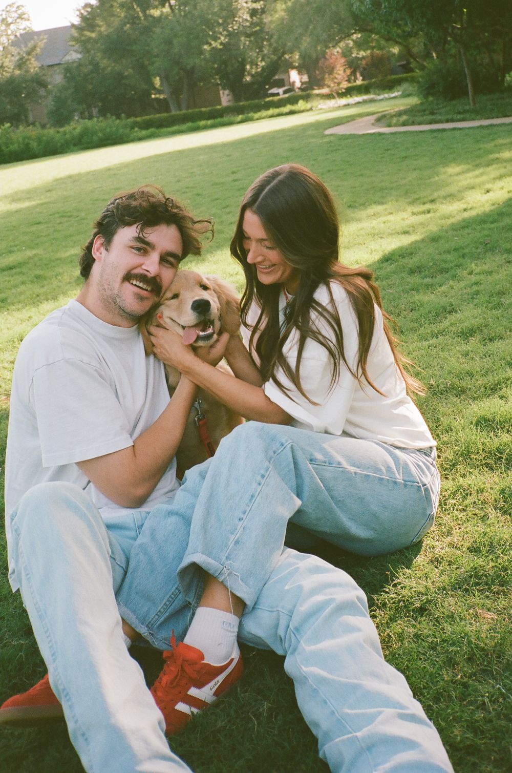 A couple in white tees and jeans sit on grass in a park, smiling and petting their golden retriever dog.