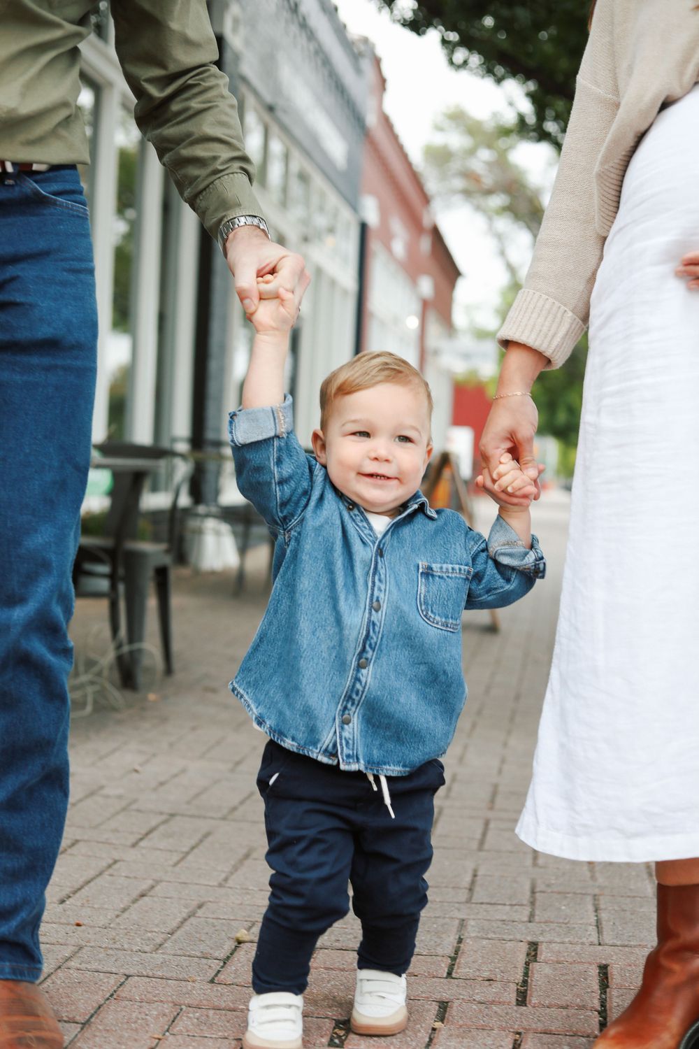Toddler in denim jacket walking while holding parents' hands on brick sidewalk.