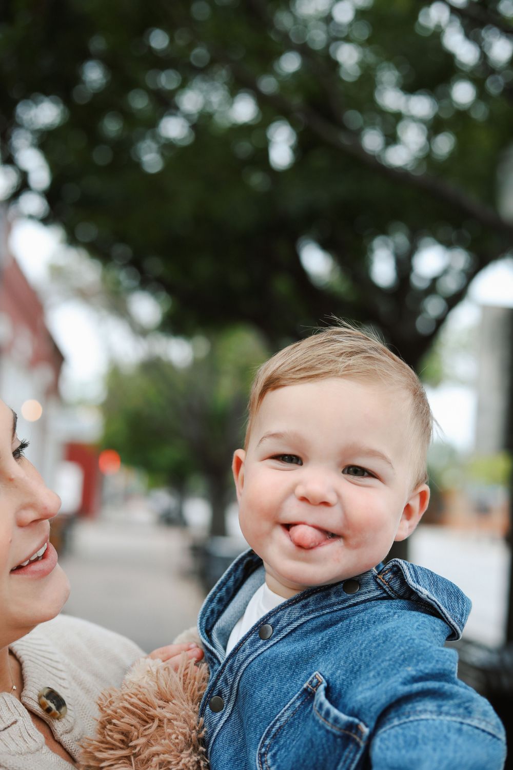 A playful denim-clad toddler being held outdoors sticks their tongue out while smiling on a sunny day.