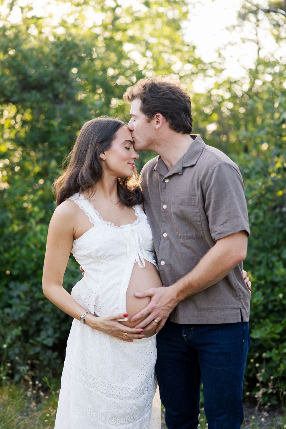 Expectant couple embracing outdoors, man kissing woman's forehead while both cradle her pregnant belly.