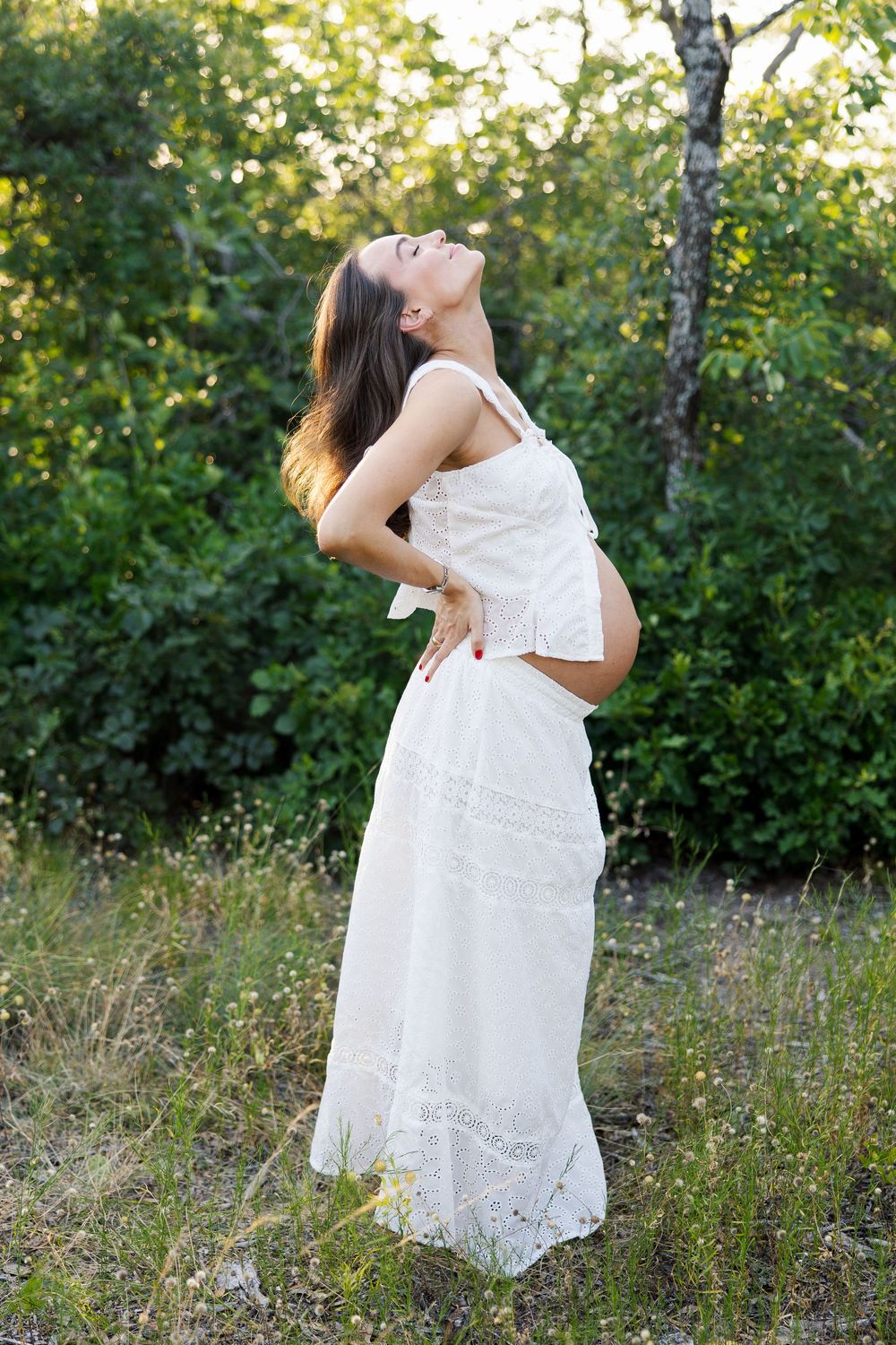 Pregnant woman in white lace dress standing outdoors among green trees, looking up with hands on her back.
