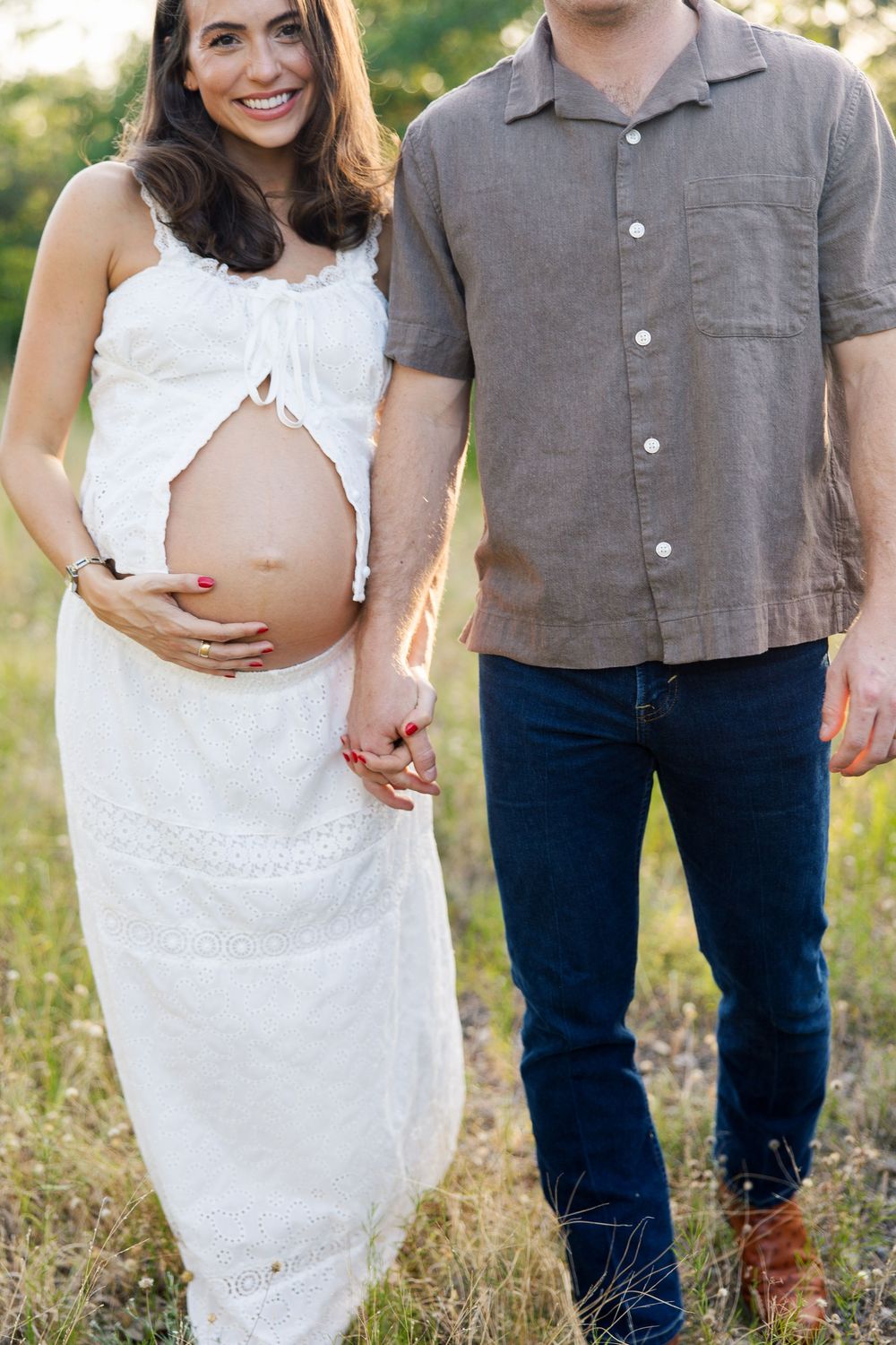 Pregnant woman in white dress holds baby bump while standing with partner in sunlit field.