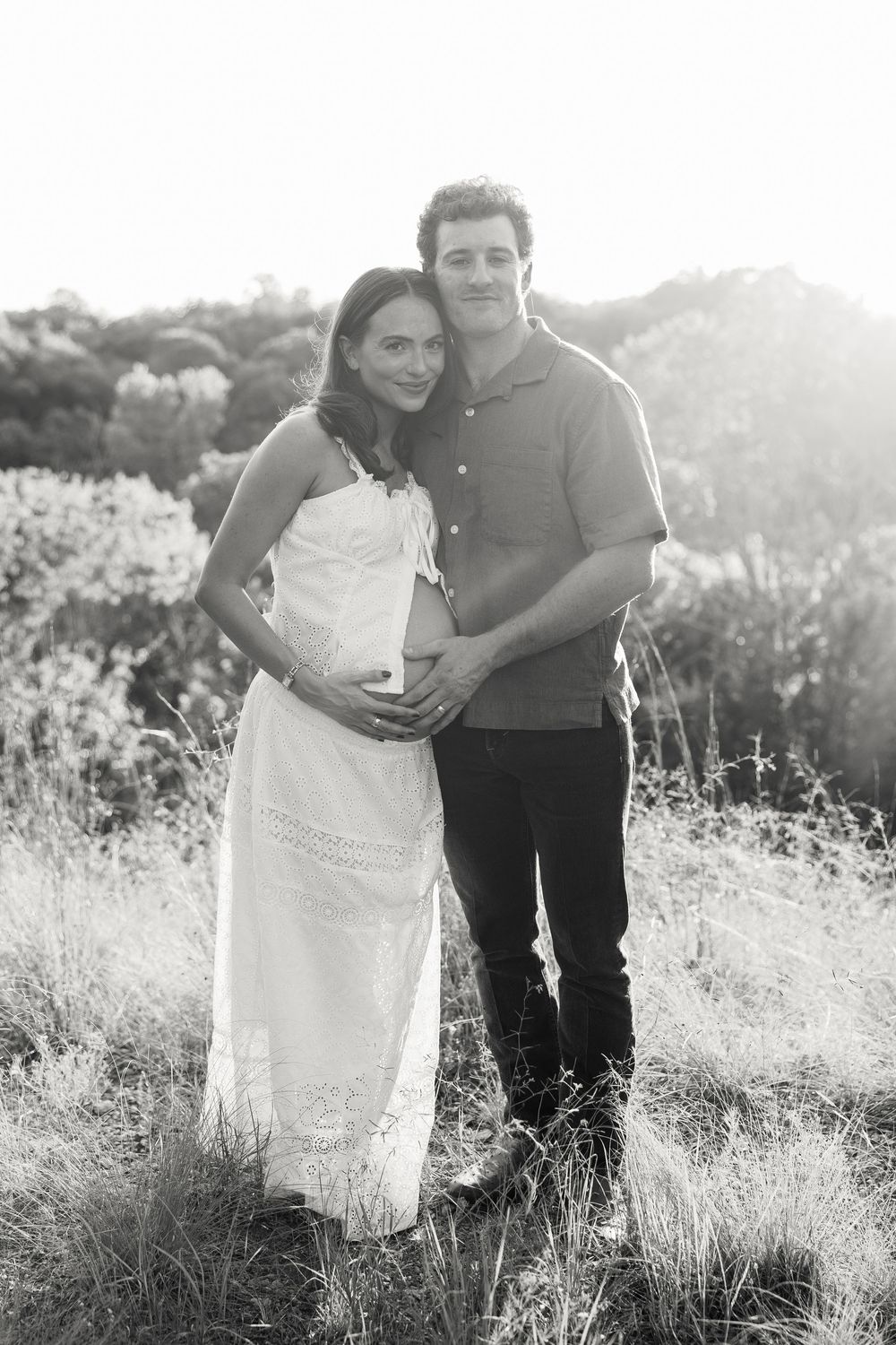 Black and white maternity photo of a couple embracing outdoors in a sunlit field, woman in white dress.