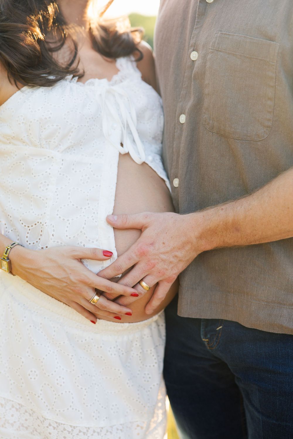 Close-up of pregnant couple holding baby bump outdoors, woman with red nails wearing gold rings.