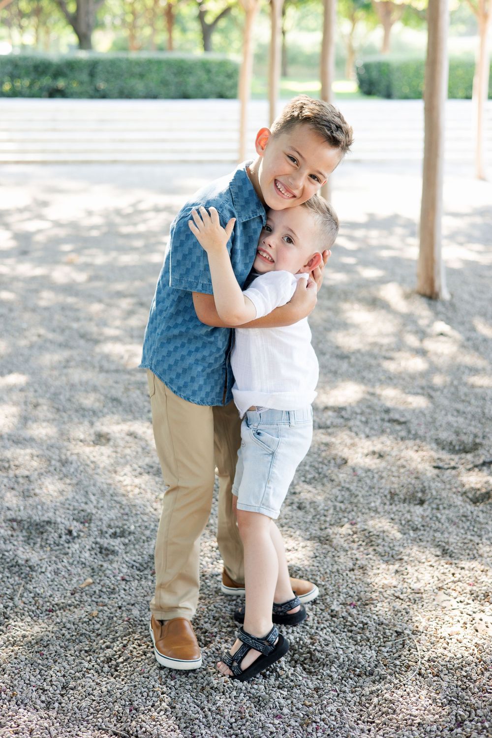 Two children share a sweet embracing moment outdoors under shaded trees.