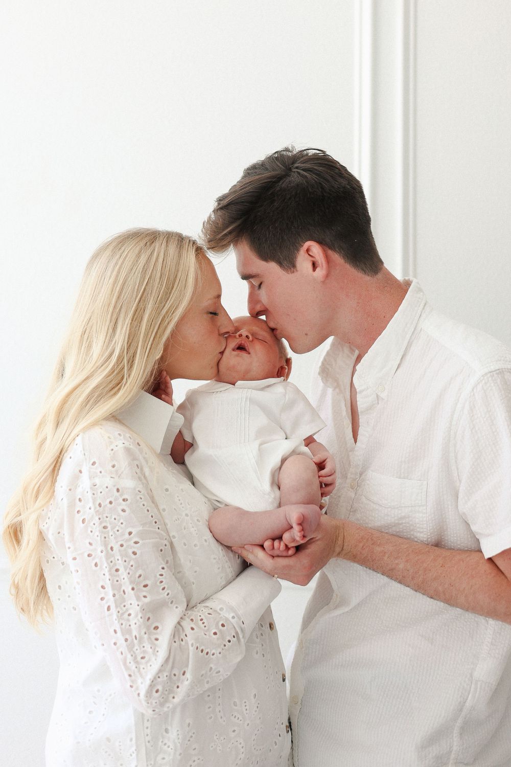 Parents tenderly kiss their newborn baby while dressed in white against a bright white background.