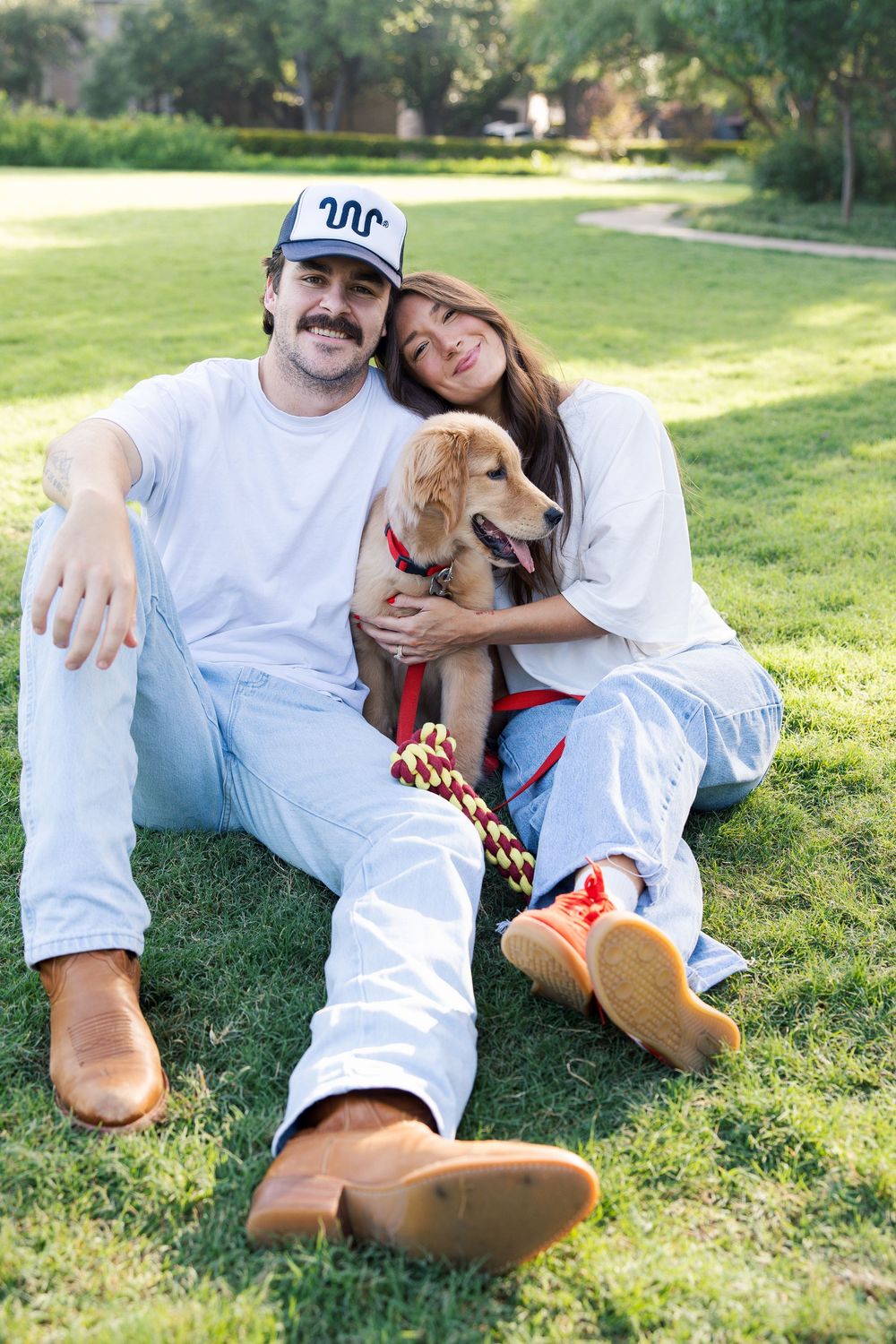 Happy couple sitting on grass in a park with their golden retriever puppy on a red leash on a sunny day.