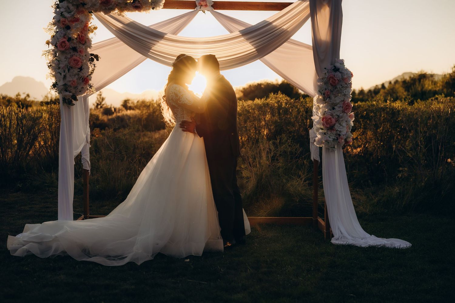 Silhouetted wedding couple stands under draped arch during golden hour sunset creating romantic backlit portrait.
