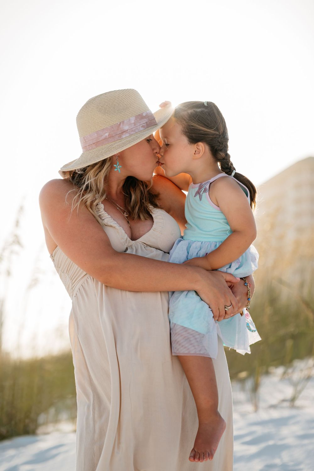Parents kiss while holding their child on a sunny beach with white sand and gentle dunes in the background.
