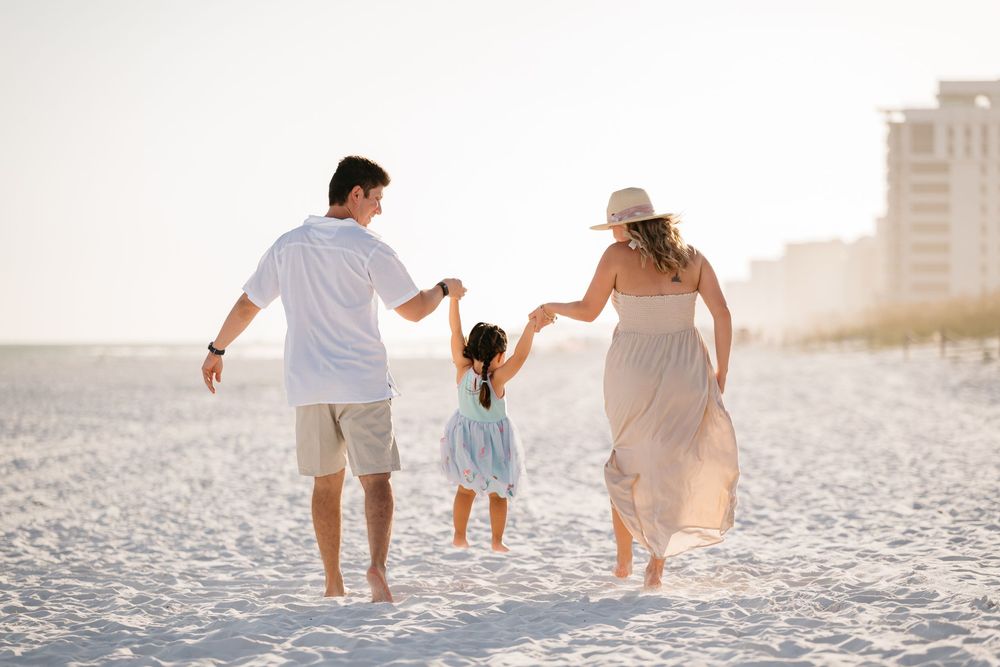 Family walking together barefoot on white sandy beach during sunset with coast cityscape in background.