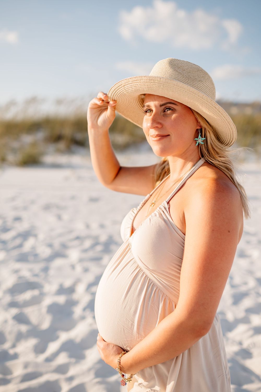 Woman in white sundress and floppy hat poses gracefully on white sandy beach at sunset.