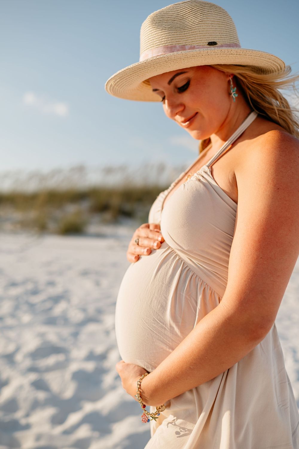 A pregnant woman in a white dress and straw hat stands on a sandy beach at sunset.