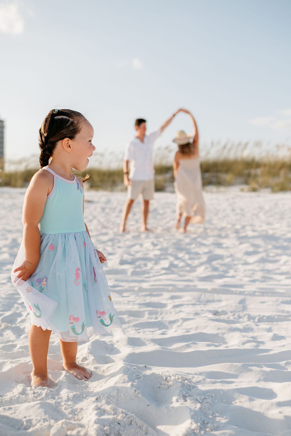 A toddler in a light blue dress plays on pristine white sand beach during a sunny summer day.