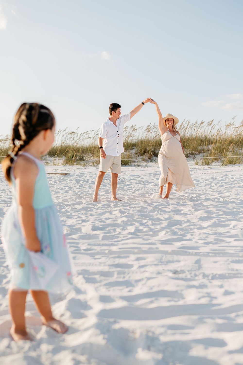 A couple takes a romantic stroll on a white sandy beach with tall grass dunes in the background.