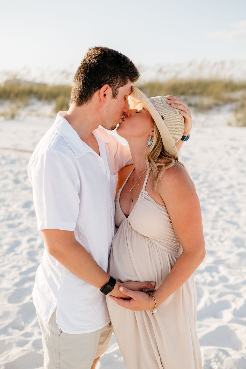 A romantic moment between a couple wearing white and beige outfits on a pristine beach at sunset.