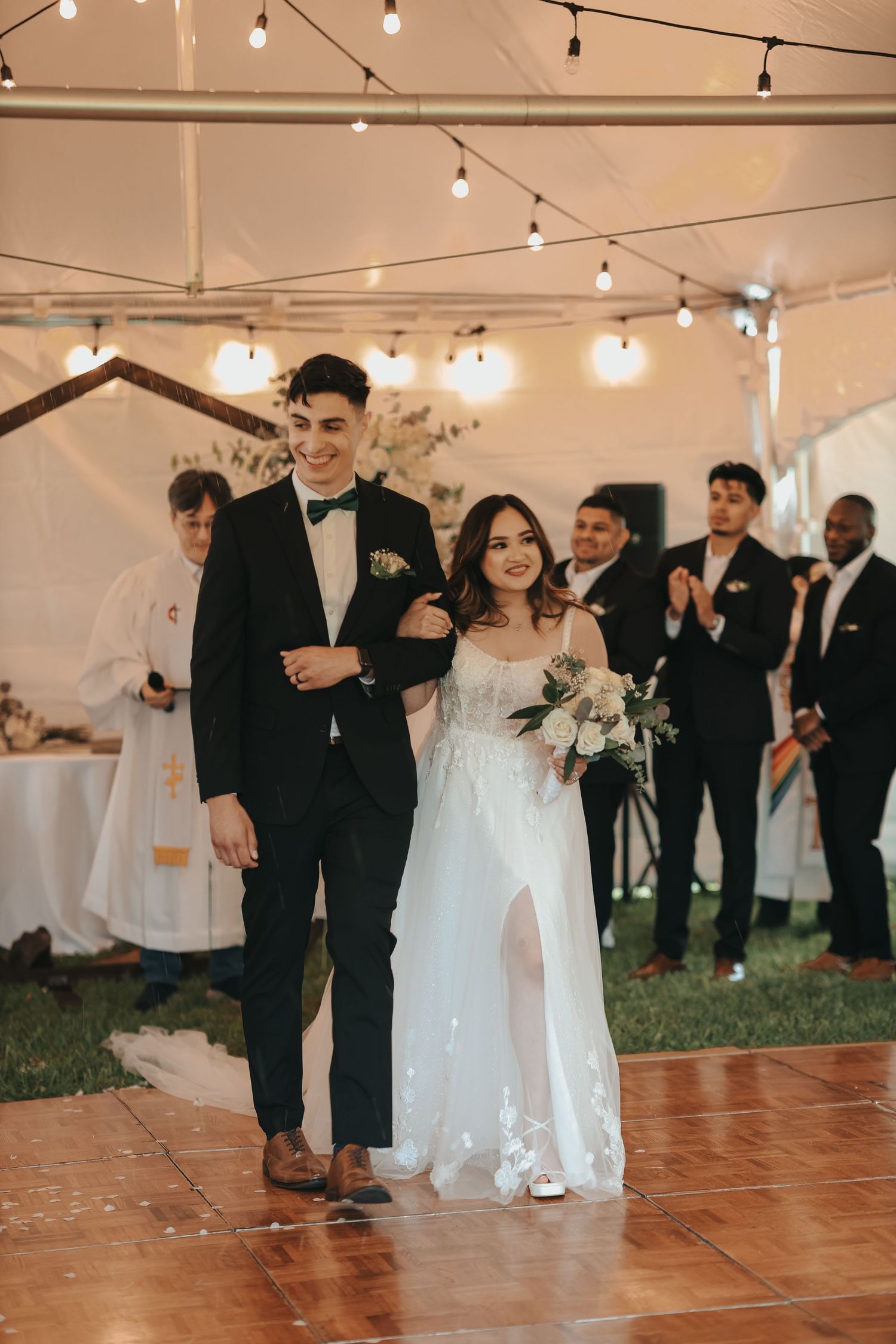 Wedding guests dance together on a wooden dance floor under string lights at an outdoor tented reception celebration.