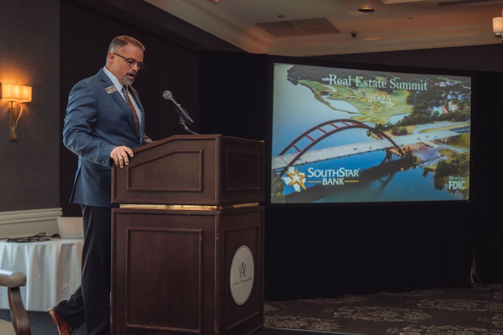 Speaker presenting at podium with projection screen showing aerial landscape view.