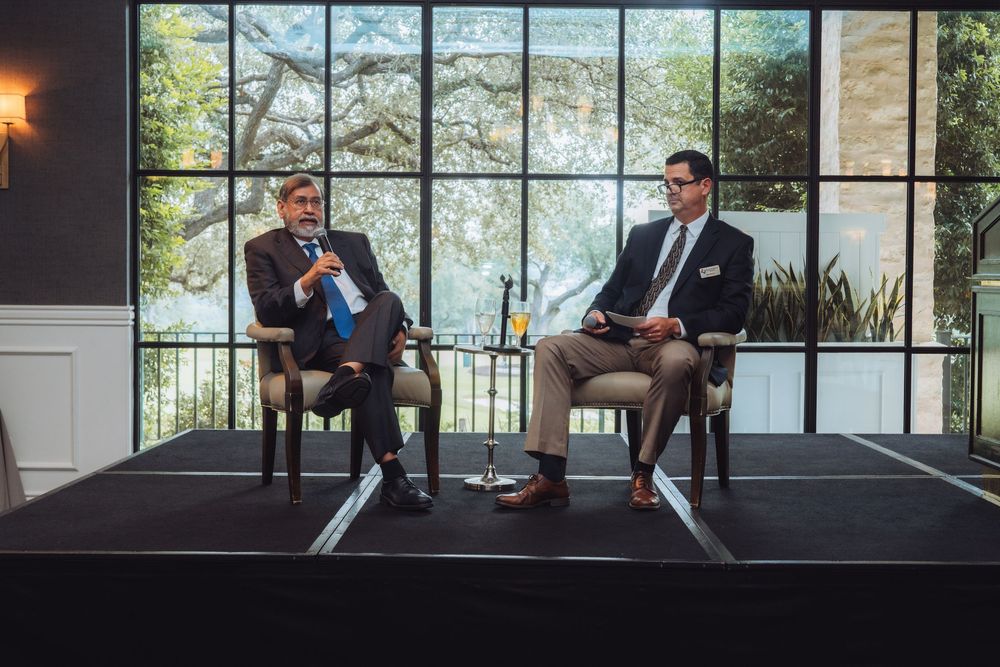 Two speakers engaged in professional discussion on stage with large windows behind them.