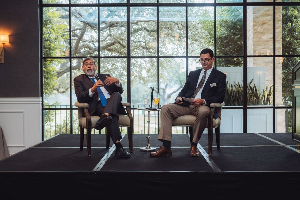Business conference speakers seated in discussion on stage with dramatic window backdrop.