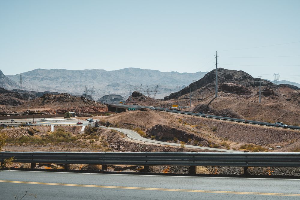 Winding desert road through rocky mountains with guard rails and power lines.