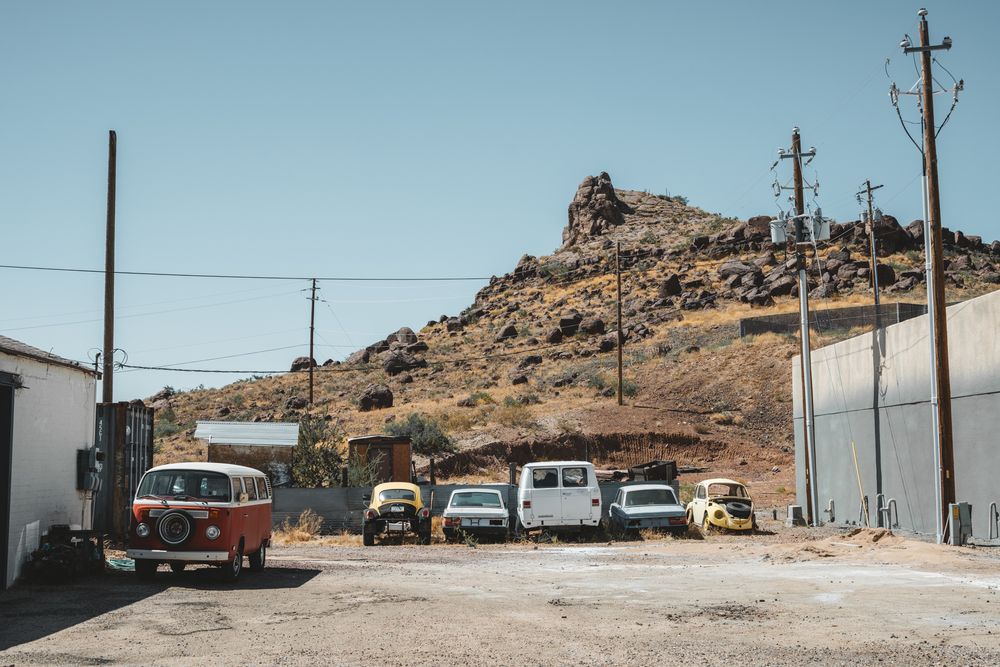 Vintage cars parked in desert lot with rocky hill and power lines in background.