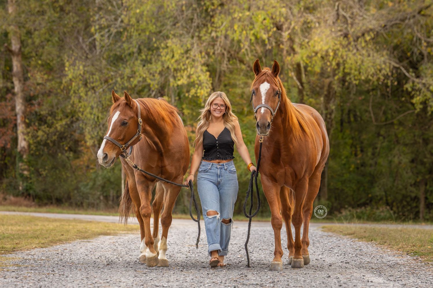 Woman in jeans and black top walking down gravel path with two chestnut horses in wooded outdoor setting.