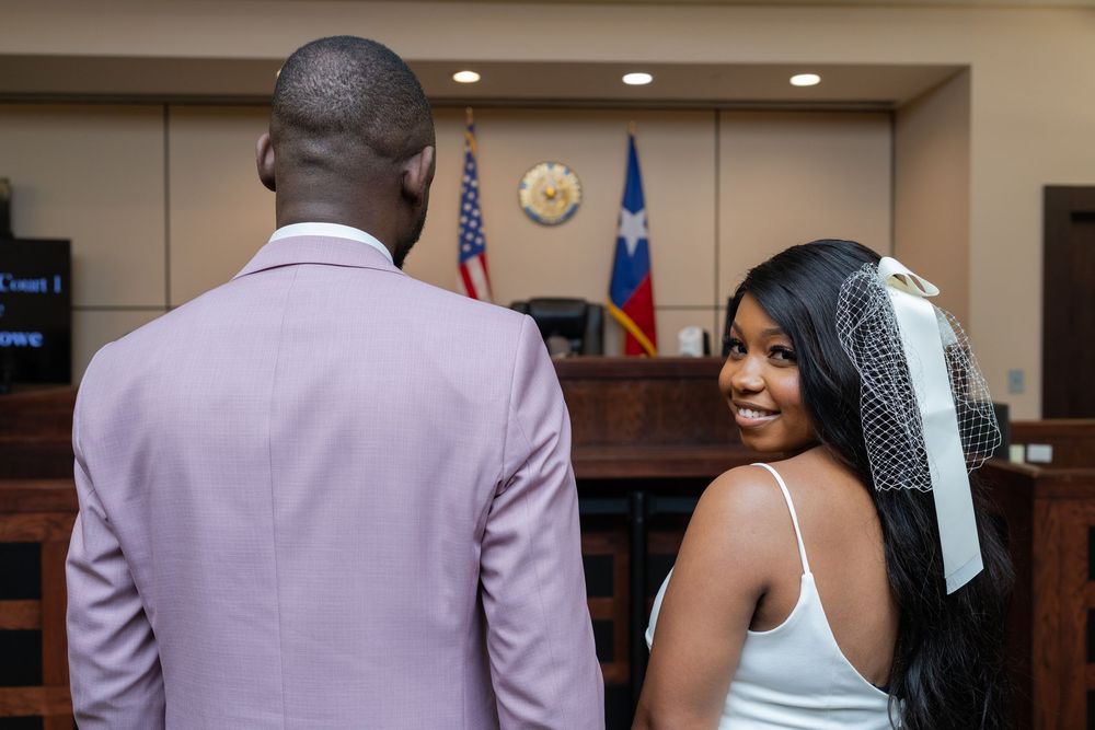A couple shares an intimate moment in a courthouse hallway wearing coordinated pink and white formal attire.