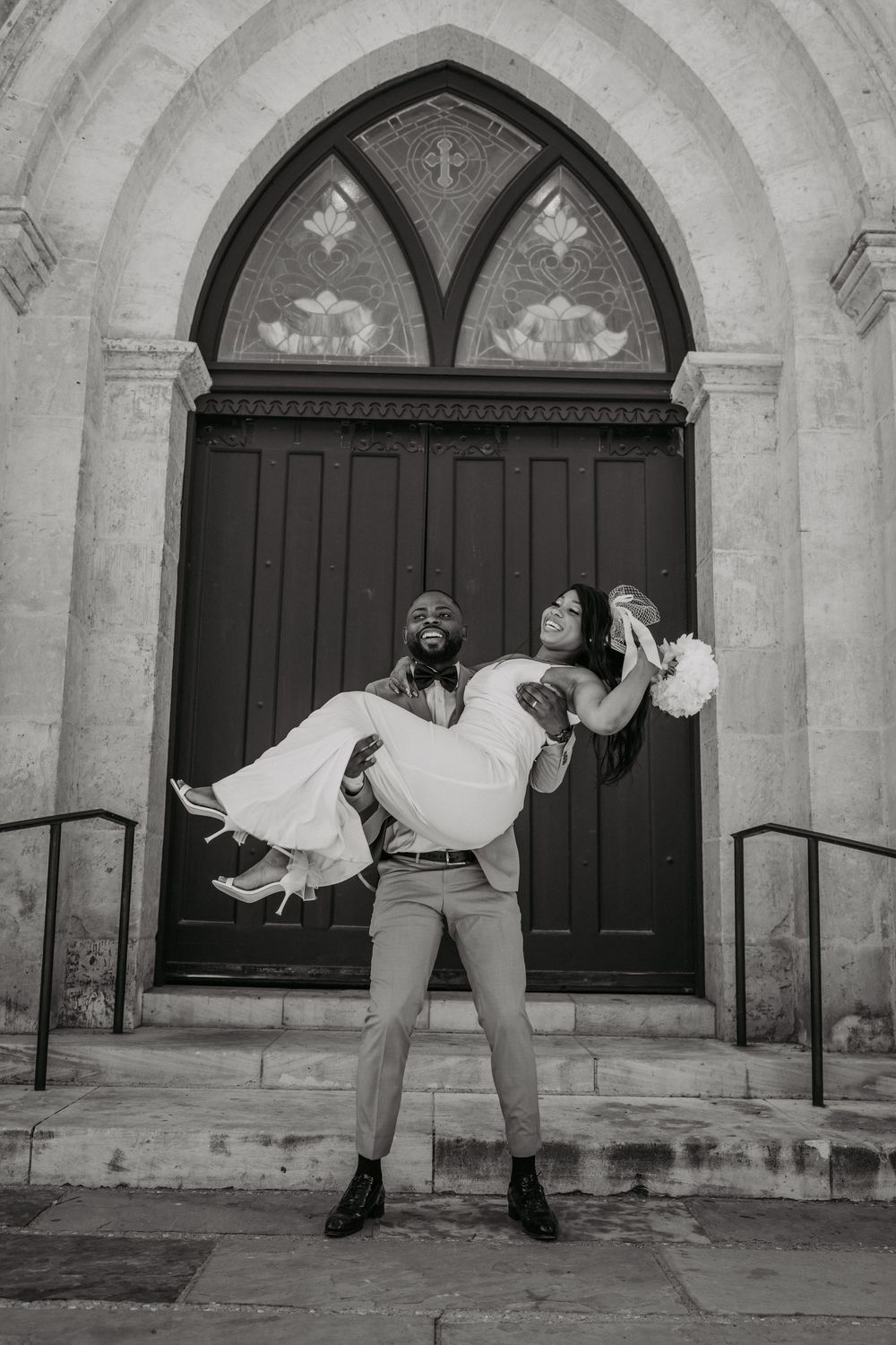 Monochrome wedding photo showing a playful moment in front of an arched church entrance.