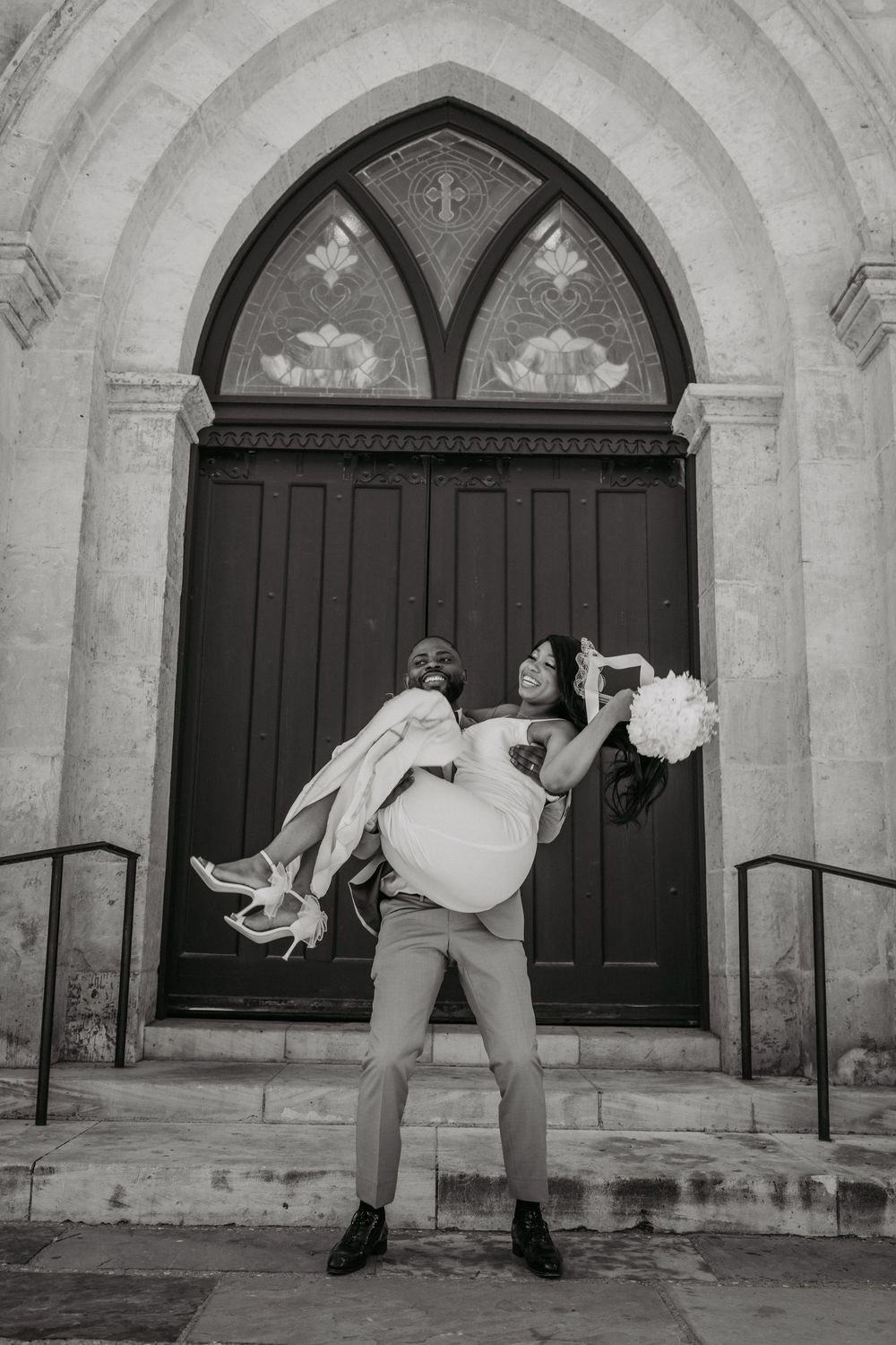 Black and white wedding photo of a couple posing playfully in front of a gothic church doorway.