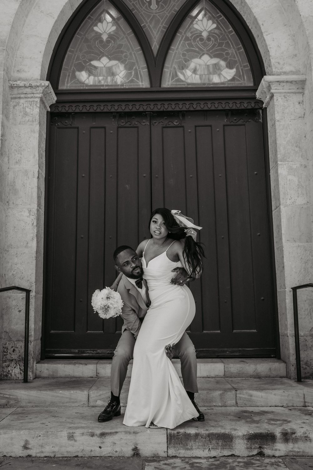 Black and white wedding portrait showing a joyful couple dancing in front of church doors.