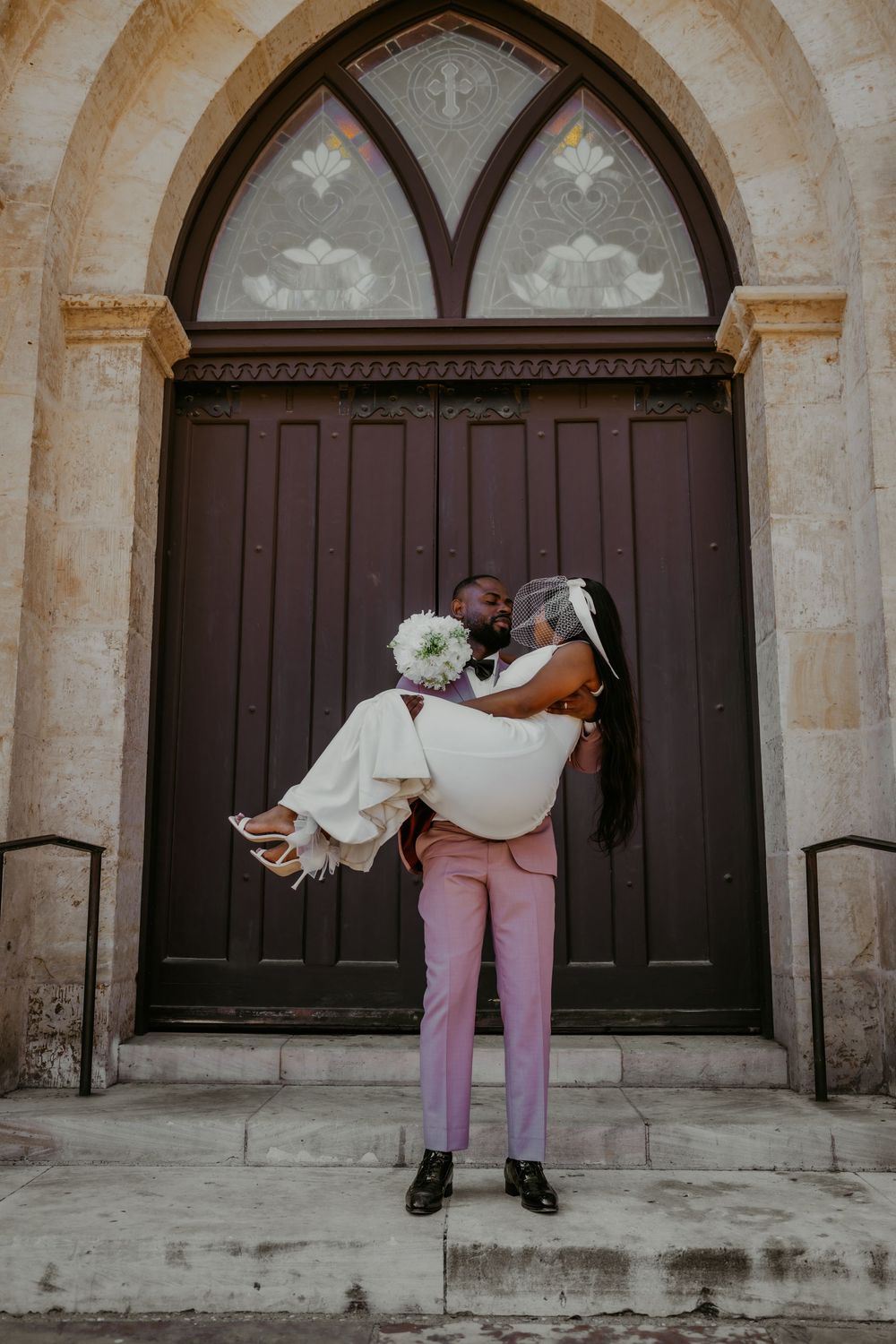 Wedding couple poses dramatically against a gothic church door with pink and white attire.