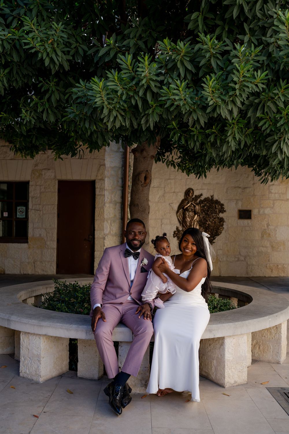 Wedding family portrait on stone bench surrounded by lush greenery.