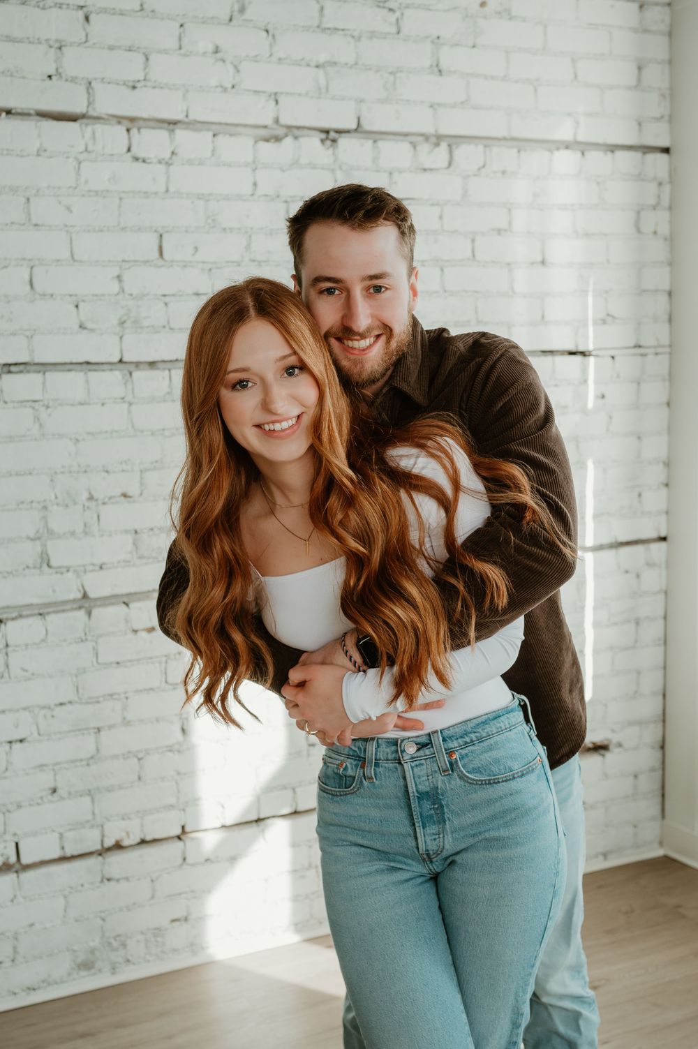 A couple in casual outfits embraces and laughs together against a white brick wall in a bright indoor space.