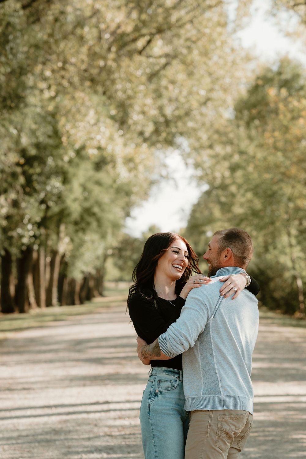 A couple shares a joyful moment while embracing on a tree-lined path during a sunny autumn day.