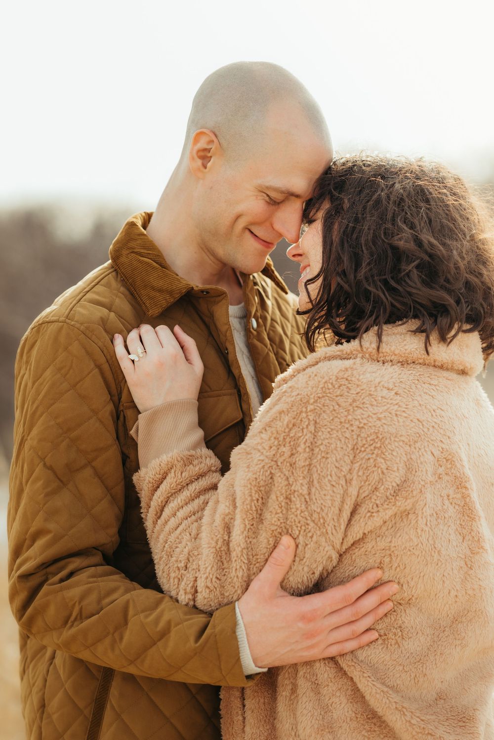 A romantic couple embraces outdoors during golden hour, wearing cozy winter clothing and sharing an intimate moment.