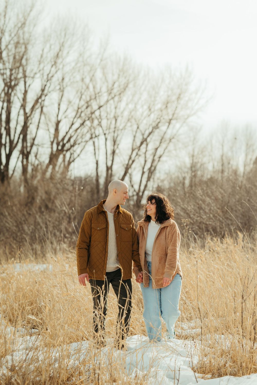 A couple walks together through tall golden grass and bare winter trees on a snowy winter day.