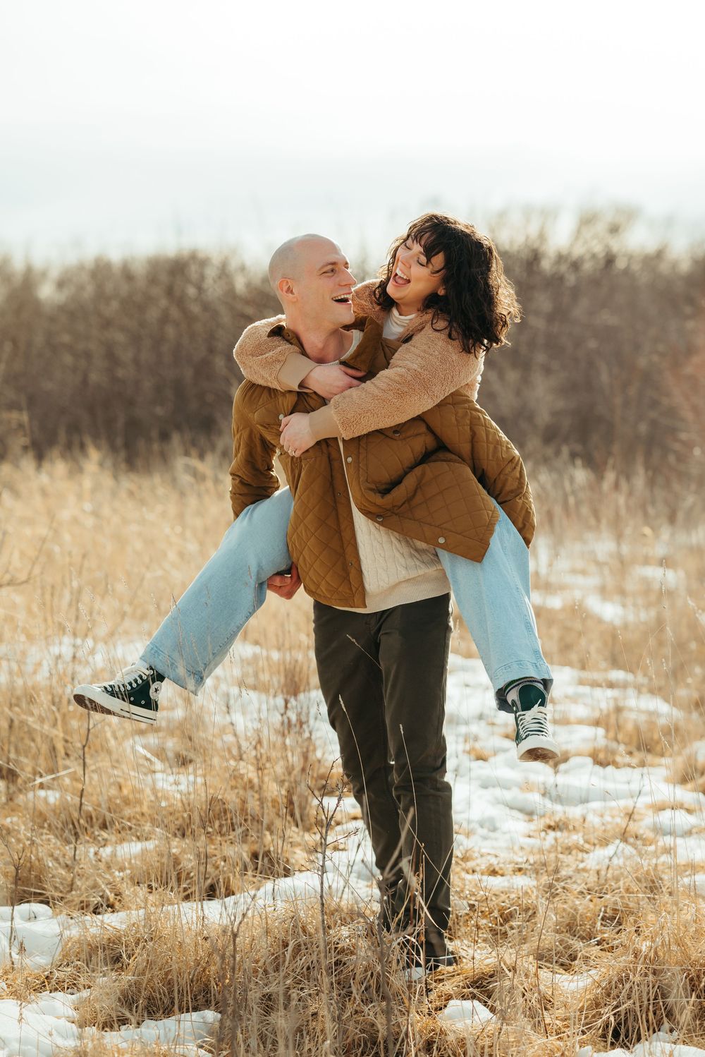 A couple embraces joyfully in a snowy winter field with dried grass and natural lighting at sunset.